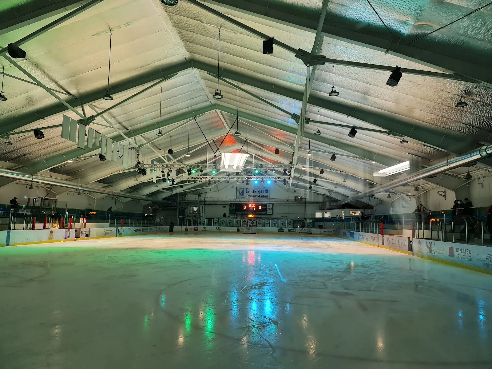 El hielo vacío bajo el techo a dos aguas, iluminado por luces de colores que se reflejan en la superficie, con el marcador en el extremo del fondo. — Centre sportif Robert-Lebel