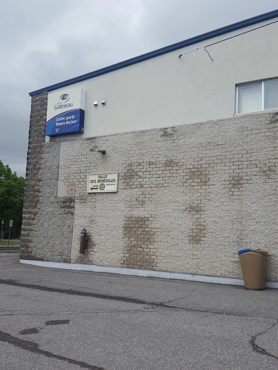 El muro lateral del Centre sportif Robert-Rochon: mampostería con revestimiento de piedra clara bajo un muro blanco con el cartel de la Ville de Gatineau que nombra el centro, un rótulo de sala en la puerta y el aparcamiento delante. — Centre sportif Robert-Rochon