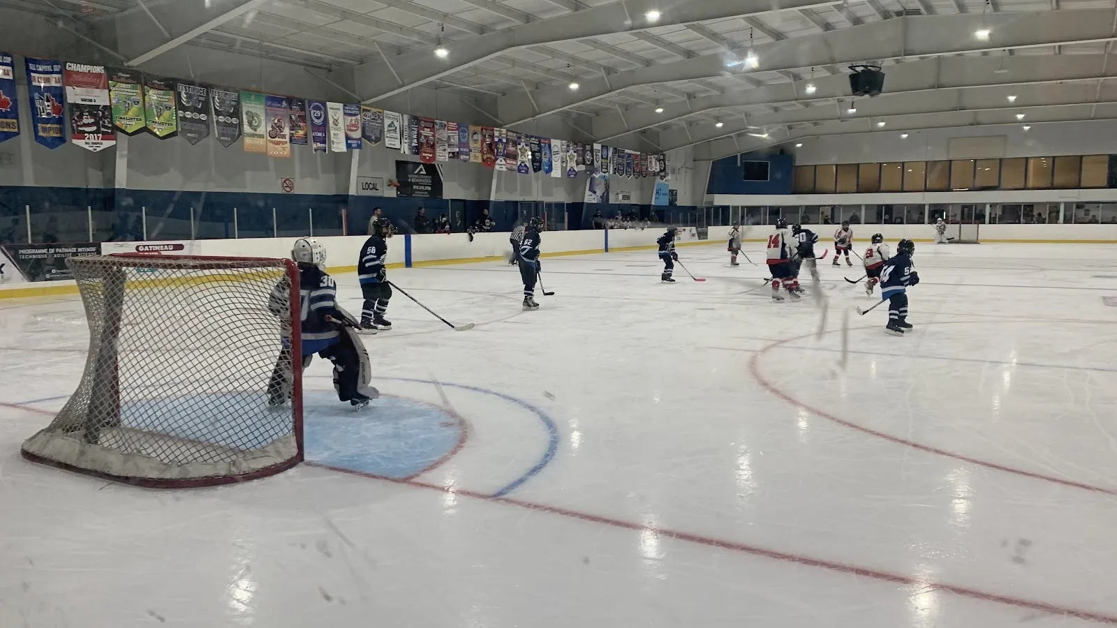 Jugadores y un portero en un extremo de una pista amplia bajo un largo techo con banderines de campeonato colgados en la pared del fondo y líneas rojas y azules sobre el hielo. — Centre sportif Robert-Rochon