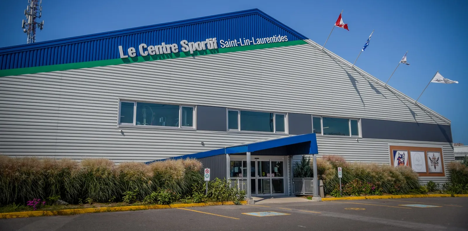 L’entrée principale de l’aréna sous une marquise bleue, avec un revêtement métallique gris, une rangée de fenêtres et le lettrage « Le Centre Sportif Saint-Lin-Laurentides » sur la bordure bleue du toit, à côté de drapeaux qui flottent au vent. — Centre sportif Saint-Lin-Laurentides