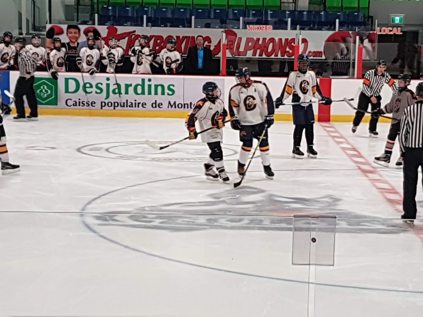 Un match à l’extrémité proche d’une grande surface de glace vu depuis les bandes, des joueurs répartis autour d’un cercle de mise au jeu, avec des panneaux de commanditaires le long du mur du fond. — Centre sportif Saint-Lin-Laurentides