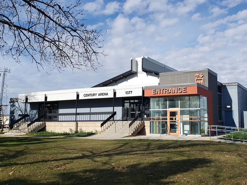 Two-tone grey building with 'CENTURY ARENA' lettering and street number '1377' above an orange-framed glass 'ENTRANCE' vestibule, bare tree branches in foreground. — Century Arena