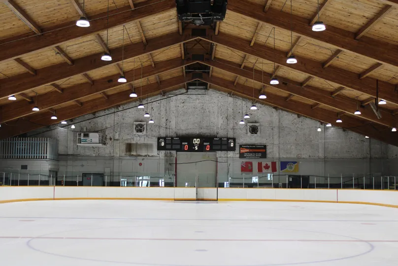Toda la pista vacía vista desde detrás de la portería, con techo a dos aguas de vigas de madera, un marcador y banderas de Canadá y de Manitoba en el muro del fondo, sin personas en el hielo. — Century Arena