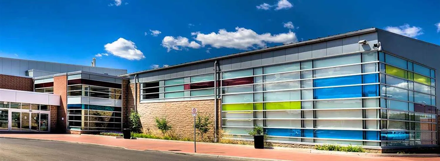 Frente del edificio del Century Gardens Recreation Centre en un día despejado: base de ladrillo y piedra con un alto muro cortina de paneles de vidrio de colores (azul, verde, guinda) arriba, una entrada de vidrio techada con puertas dobles a la izquierda y un acceso pavimentado con áreas verdes jóvenes. — Century Gardens Recreation Centre