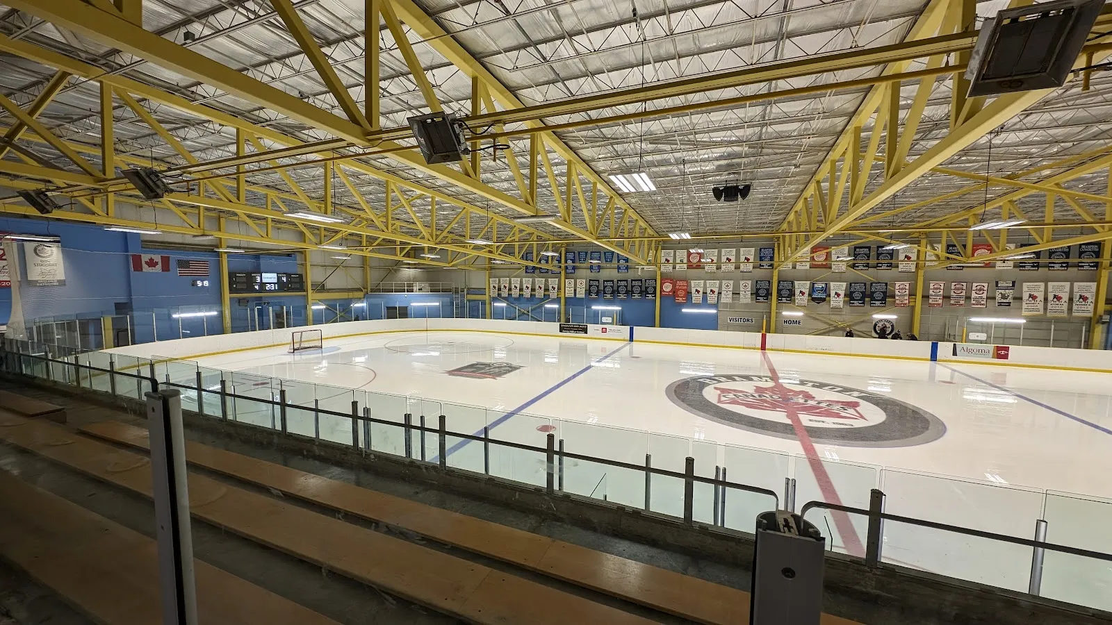 Toda la pista dentro de una arena con armaduras amarillas en el techo, una bandera de Canadá y una de Estados Unidos en la pared del fondo, mantas de campeonato a lo largo de ambas paredes laterales y un gran logotipo rojo y blanco de las "Brampton Canadettes" en el centro de la pista; dos personas de pie junto a la pista muy al fondo. — Century Gardens Recreation Centre