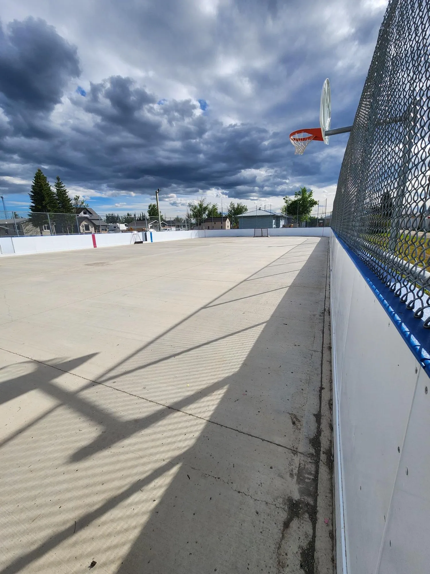La pista exterior de Champion sin hielo, vista a lo largo de las vallas, con una cerca de malla, un aro de baloncesto y un cielo nublado. — Champion Outdoor Arena