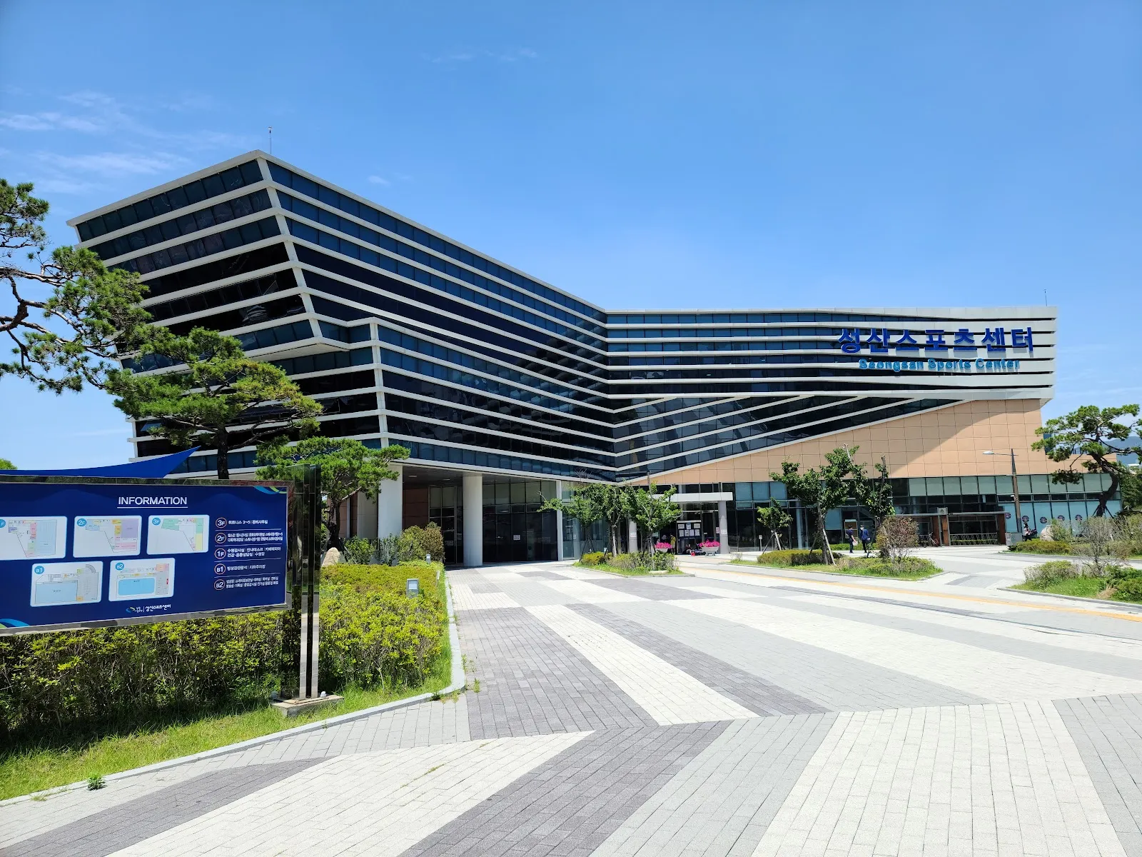 The angular glass-and-stone Seongsan Sports Center building with its blue name sign, seen across the paved plaza on a clear day. — Seongsan Sports Center Ice Rink (Changwon)