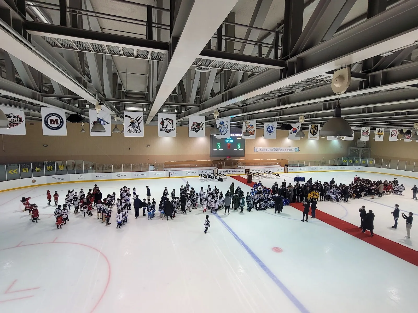 A crowd of skaters gathered along the boards on the ice of the Seongsan Sports Center rink in Changwon, seen across the hall under the steel roof. — Seongsan Sports Center Ice Rink (Changwon)