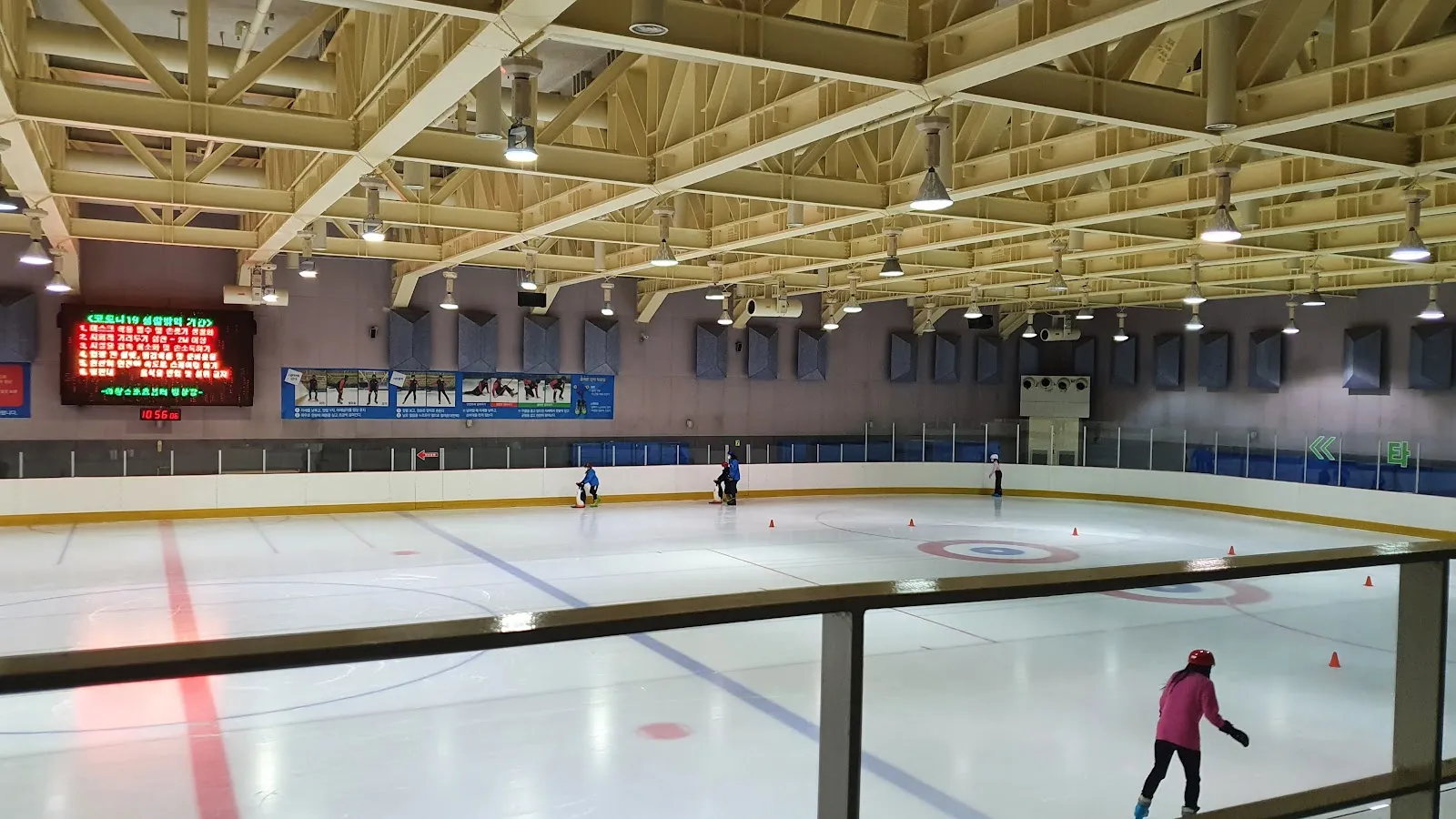 발코니 난간에서 본 Uichang Sports Center의 아이스링크. 목재 천장, 전광판, 펜스가 넓은 빙판을 둘러싸고 있으며 스케이터 몇 명과 콘이 보여요. — Uichang Sports Center Ice Rink (Changwon)