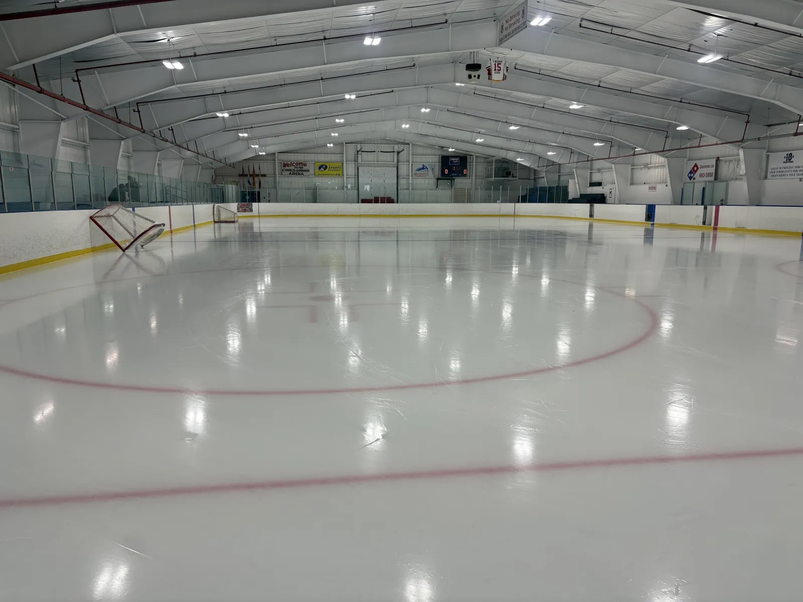 The empty ice seen from centre ice, with the far boards and glass under a white arched roof with rows of lights. — Charles Gorman Arena