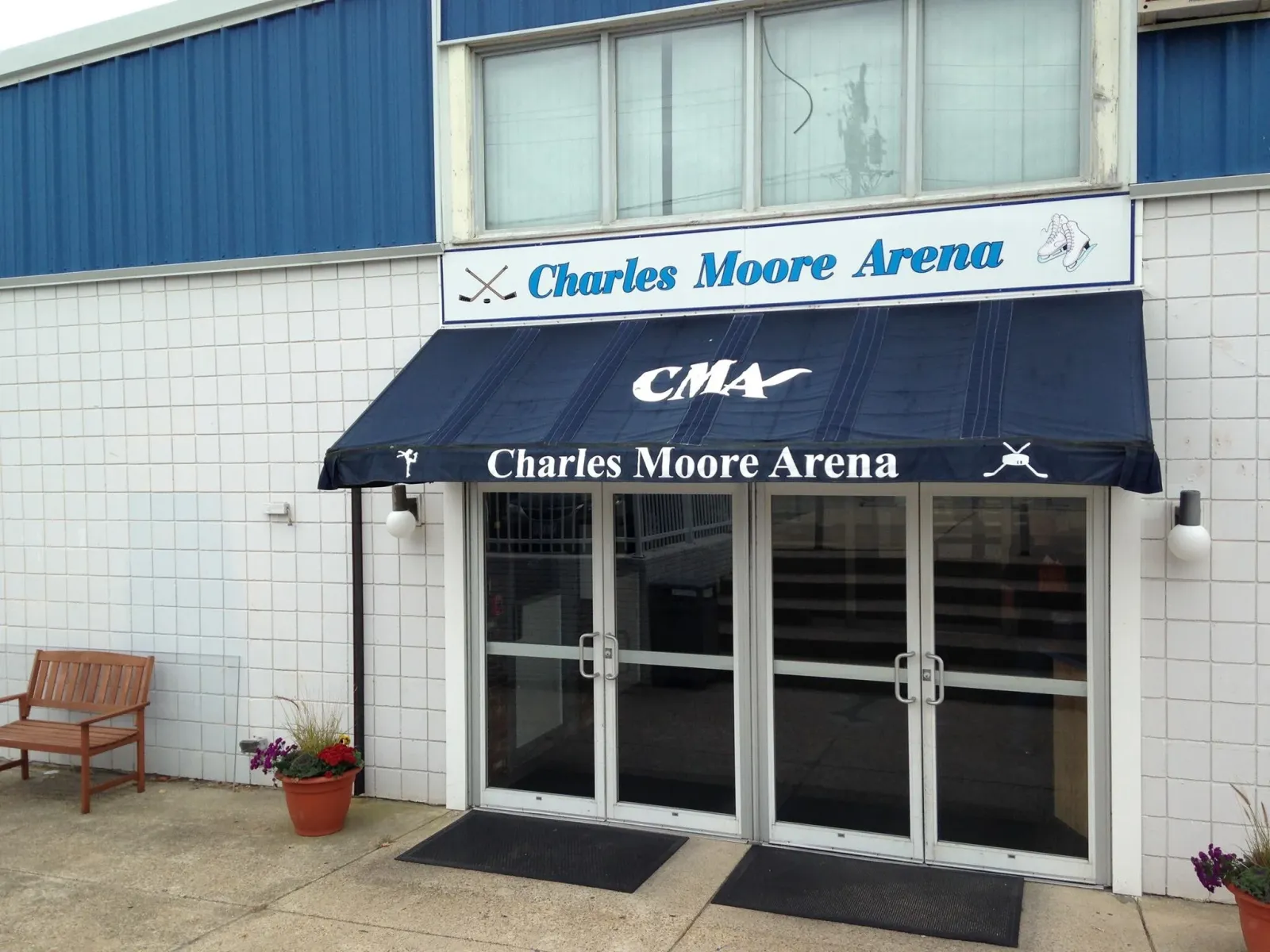 The arena entrance under a navy "CMA" awning, with a blue "Charles Moore Arena" sign on the white block wall above the glass doors. — Charles Moore Arena