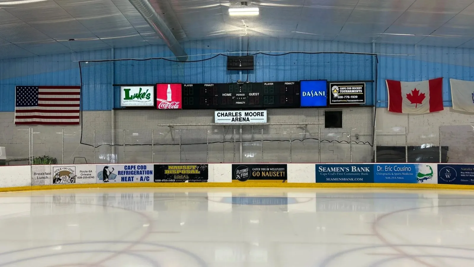 The empty rink under a blue-trussed roof, with US and Canadian flags, a scoreboard and sponsor boards along the far end. — Charles Moore Arena