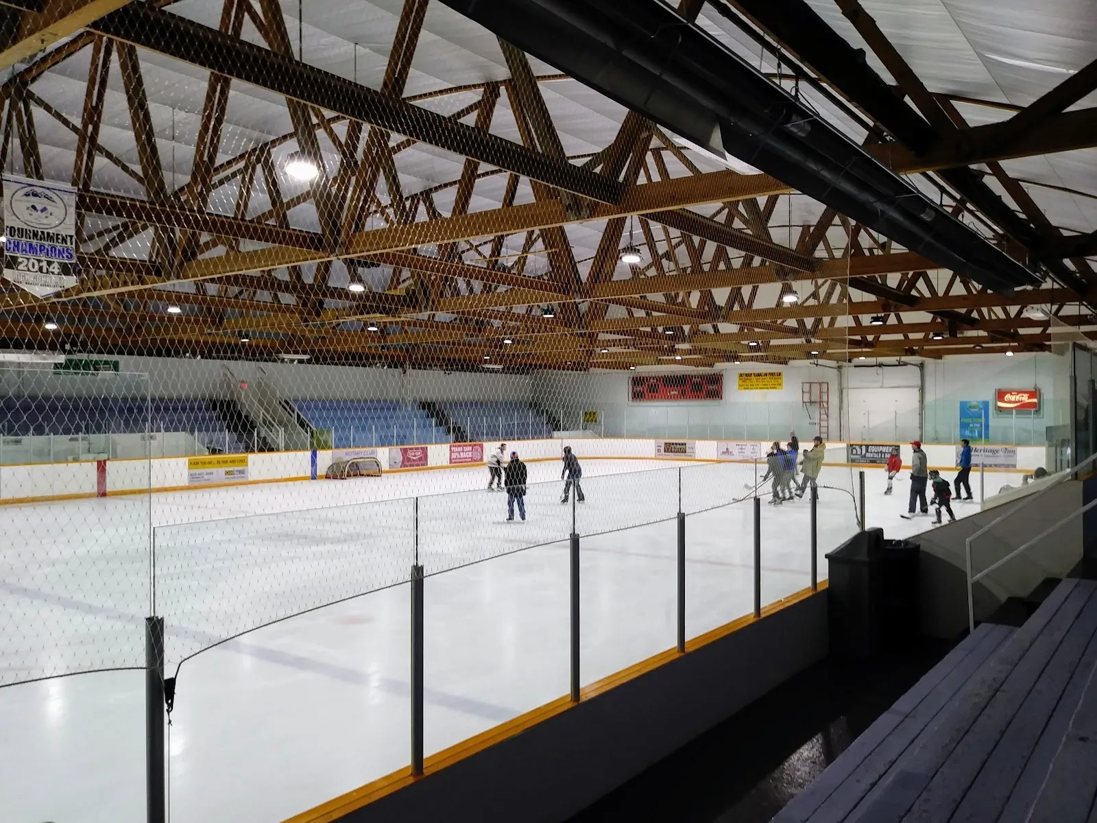 A few skaters on the ice under the exposed timber roof trusses, seen over the netting from the wooden bleachers. — Charlie Cheesman Memorial Ice Arena