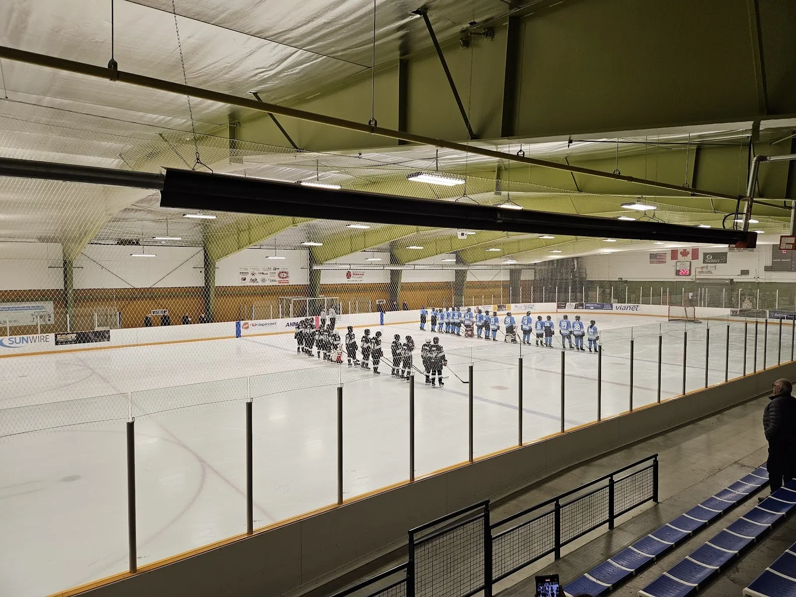 A wide view across the ice at Chelmsford Community Centre and Arena from the stands, with two teams lined up along the far boards before a game and flags and sponsor signs on the end wall. — Chelmsford Community Centre and Arena