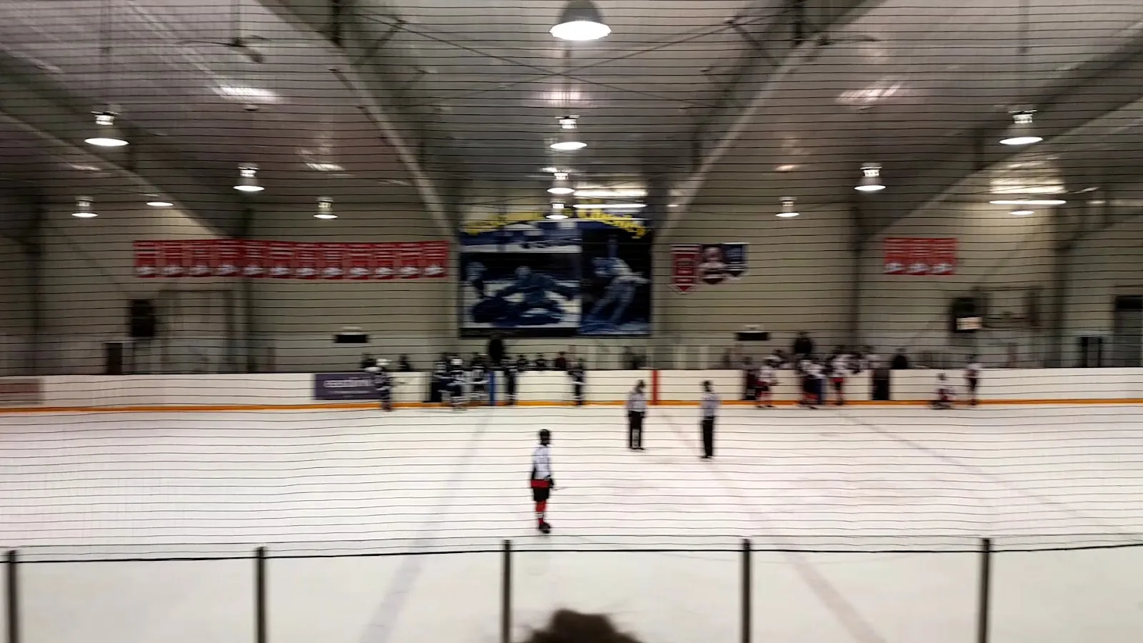 The ice seen from the stands during a game, with a red-and-white banner row and a video board on the far wall. — Chesley Community Centre