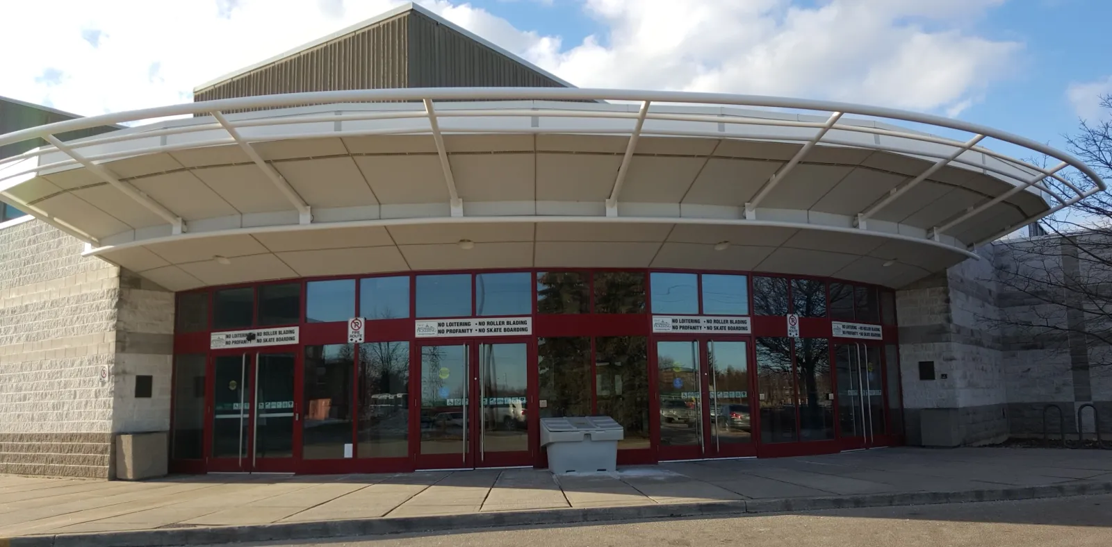 El toldo curvo sobre la entrada acristalada de marco rojo de la arena del Chestnut Hill Developments Recreation Complex, bajo un cielo azul claro. — Chestnut Hill Developments Recreation Complex (Pickering)