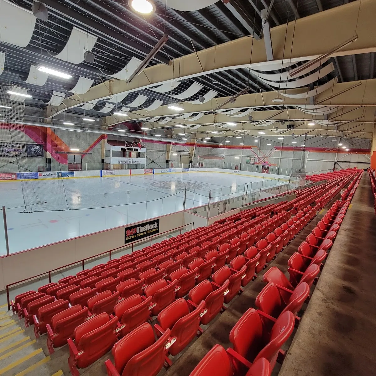 Una de las pistas del complejo desde lo alto de los asientos rojos: el hielo, las vallas del extremo y un panel acústico blanco y curvo en el techo. — Chestnut Hill Developments Recreation Complex (Pickering)