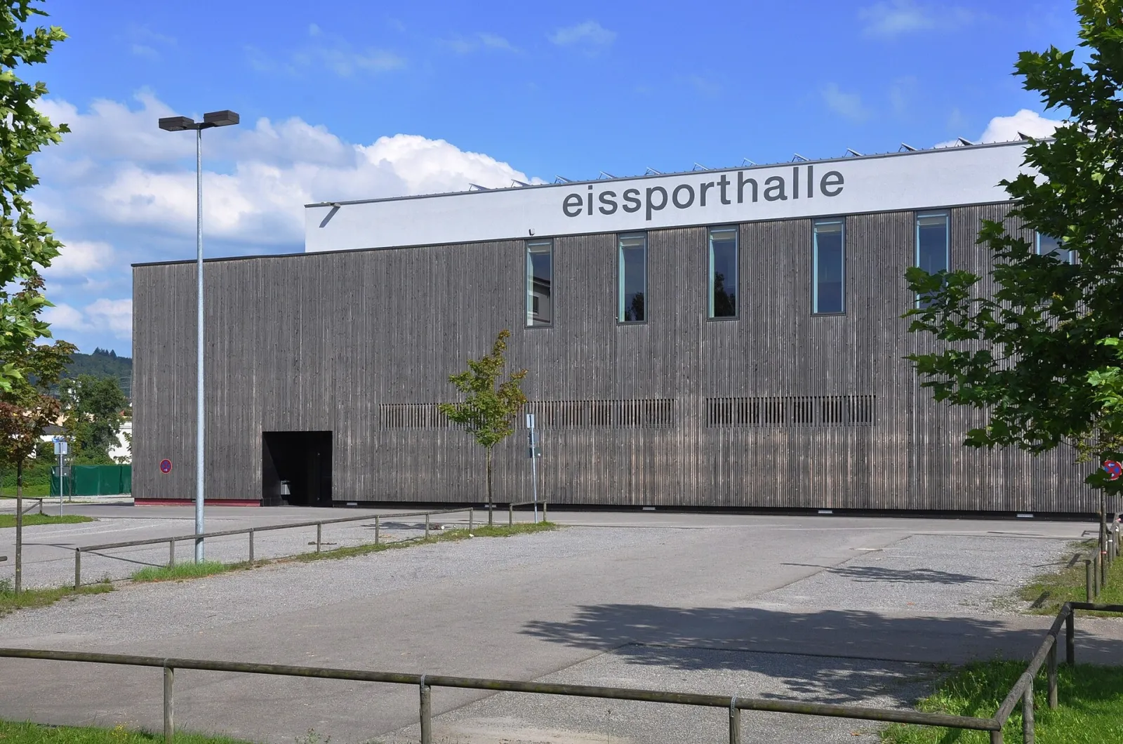 The long timber-clad side of the Ravensburg Eissporthalle with its lettering on the roofline, seen from the car park. — CHG Arena Ravensburg