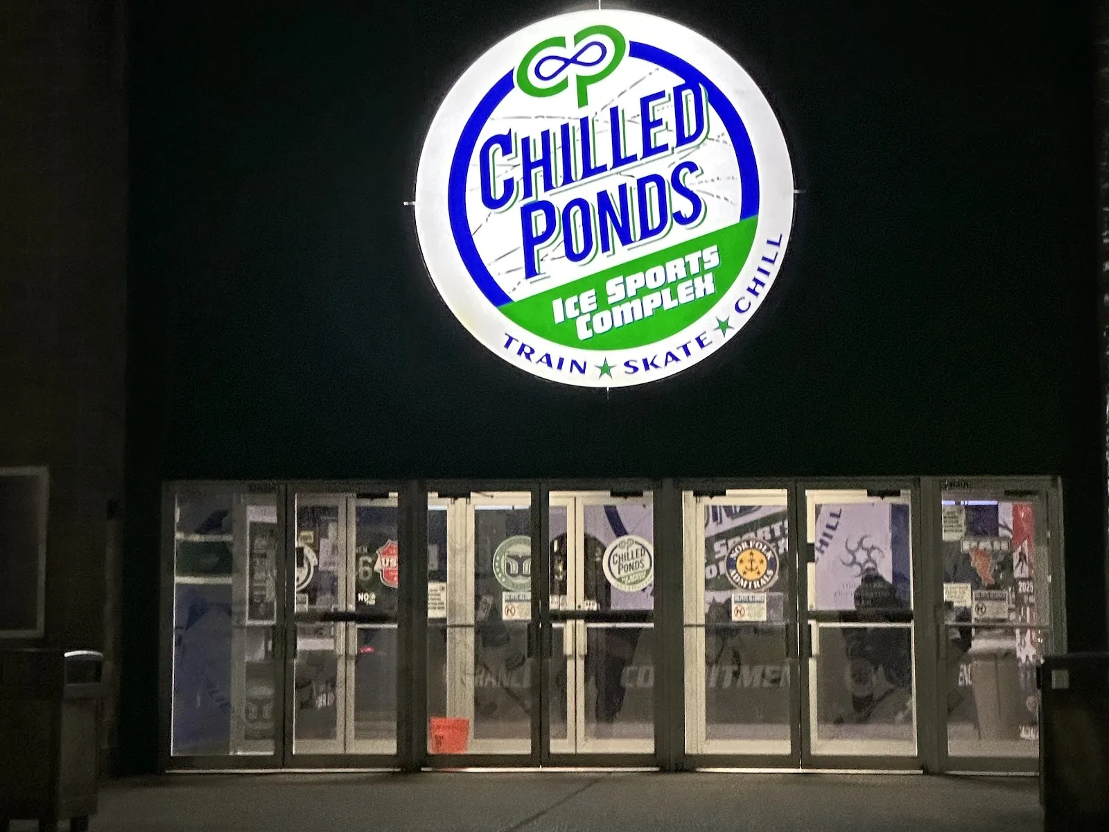 Illuminated circular "Chilled Ponds Ice Sports Complex" sign above the glass entrance doors, photographed at night. — Chilled Ponds Ice Sports Complex