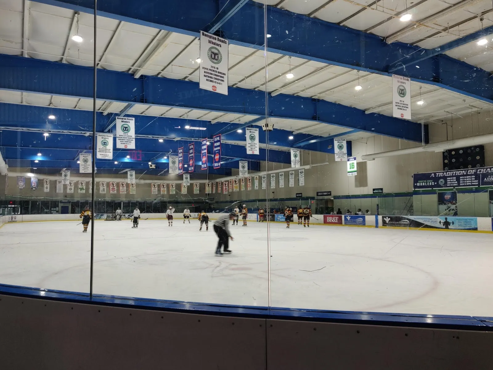 A wide sheet seen through the glass, a lone skater far down the ice under a blue-trimmed roof with banners hung along the far wall. — Chilled Ponds Ice Sports Complex