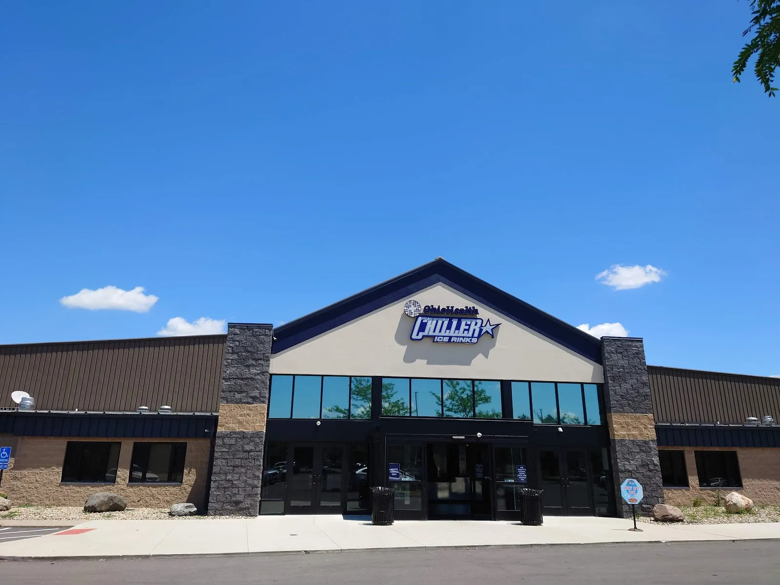 Front entrance of the building on a clear day: dark metal-and-stone facade with the illuminated "OhioHealth Chiller Ice Rinks" sign over the glass entry doors, empty sidewalk and parking lot in front. — OhioHealth Chiller Dublin