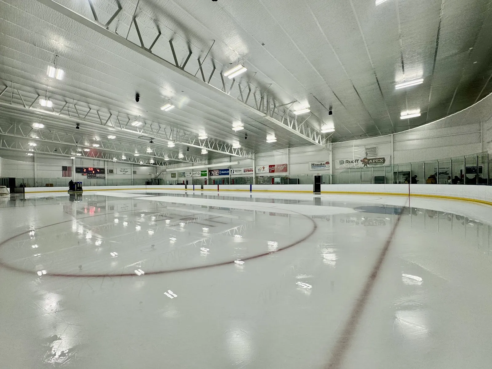 Empty NHL-sized ice sheet from ice level: scoreboard reading 0:00, American flag, sponsor banners (Honda, OhioHealth, Ruoff Mortgage) along the boards, arched white roof trusses overhead. No skaters on the ice. — OhioHealth Chiller Dublin