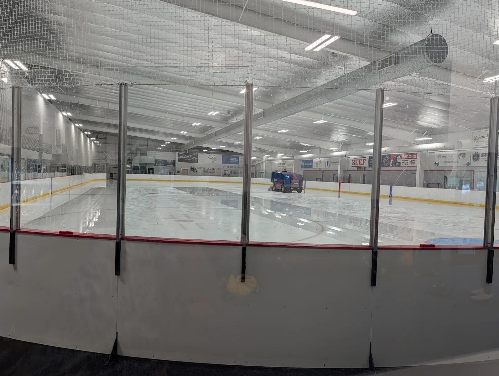 The empty ice being resurfaced, seen through the glass from behind the boards, with New Albany Hockey banners on the far wall. — OhioHealth Chiller Easton