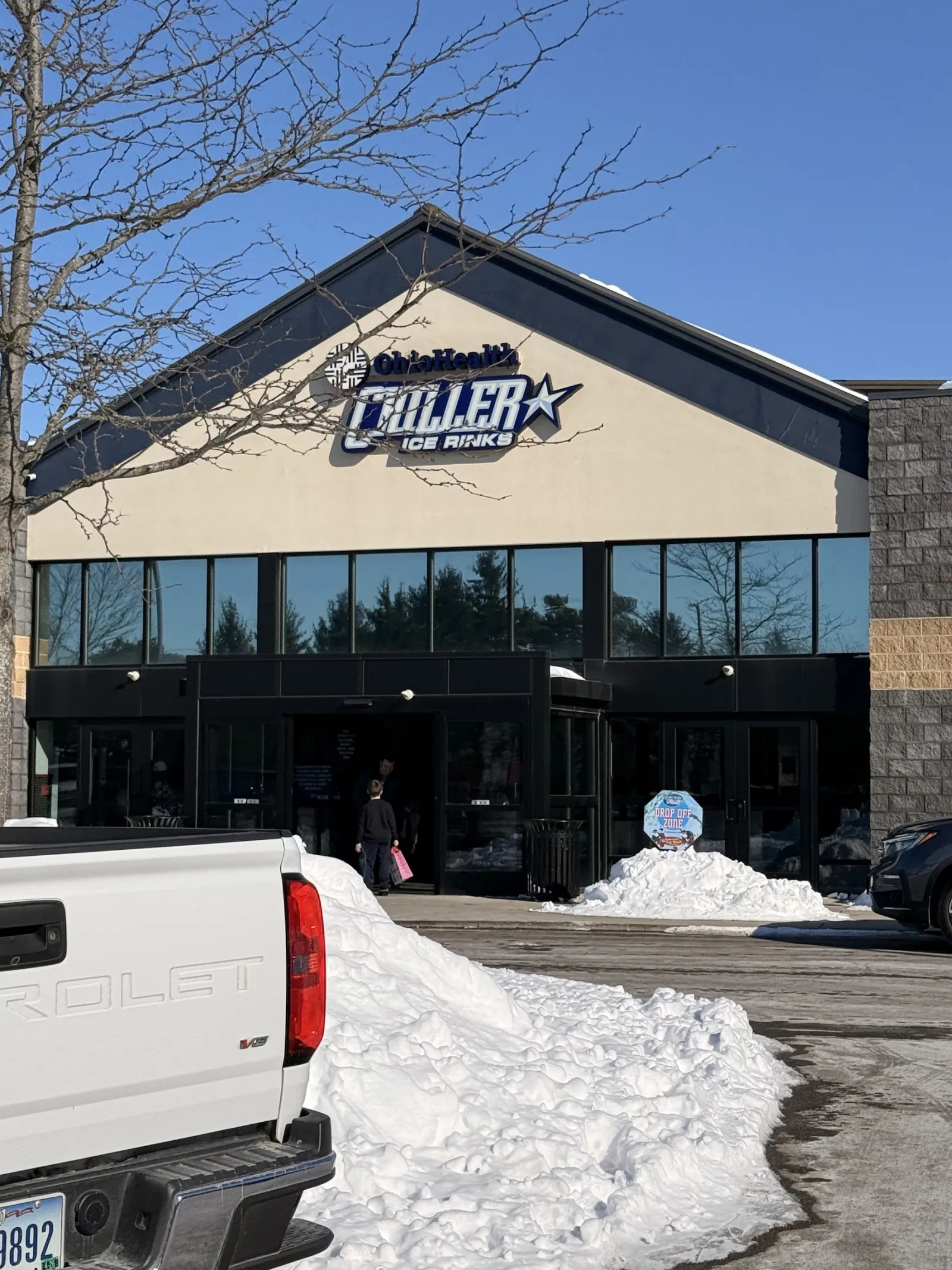 The front of the building on a sunny winter day, with the OhioHealth Chiller Ice Rinks sign above a glass entrance and snowbanks in the lot. — OhioHealth Chiller Ice Works
