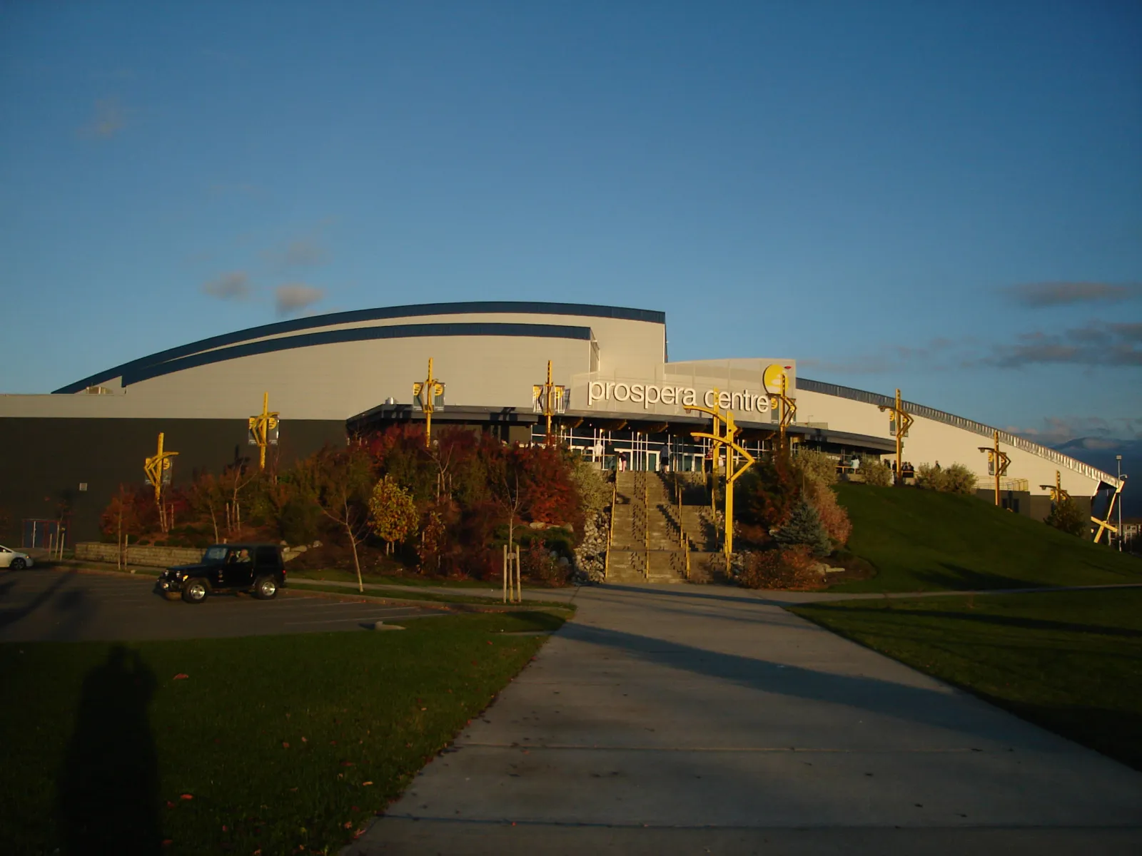 The arena at sunset under its former name, with prospera centre lettering over the glass entrance, a curved roofline, front steps and landscaping. — Chilliwack Coliseum