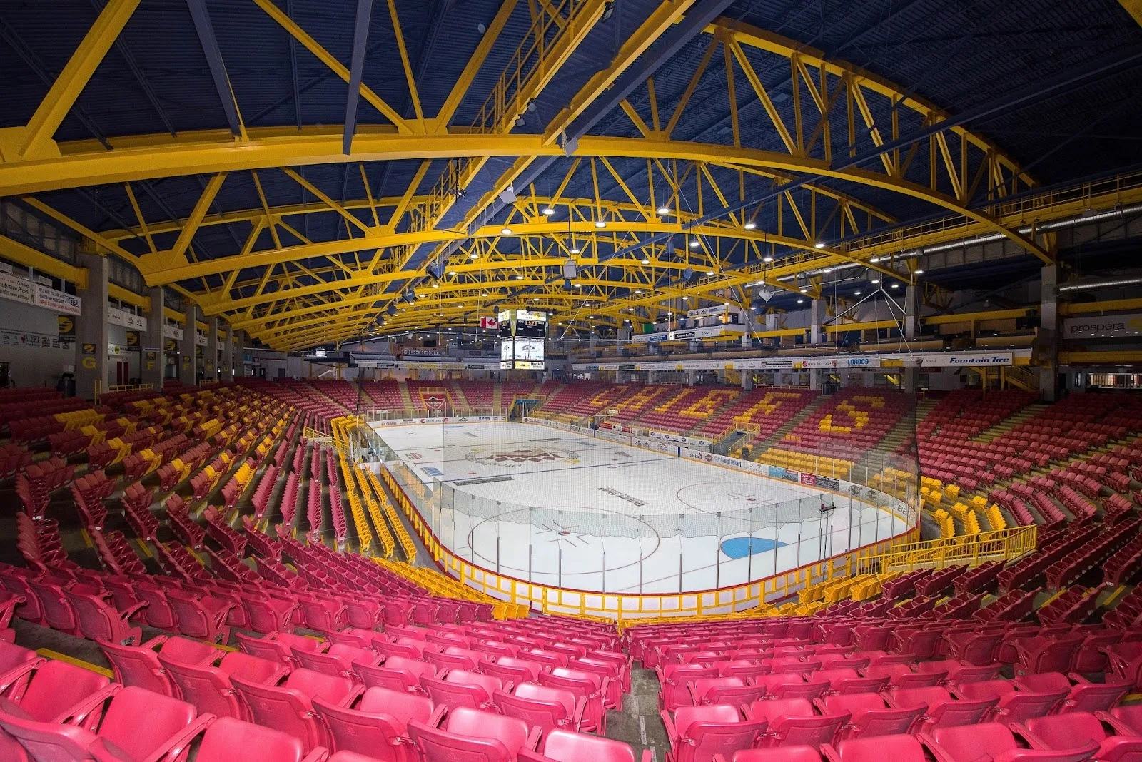 The coliseum from the end, red seats banked round the sheet and a yellow-trussed ceiling above a pale ice surface. — Chilliwack Coliseum