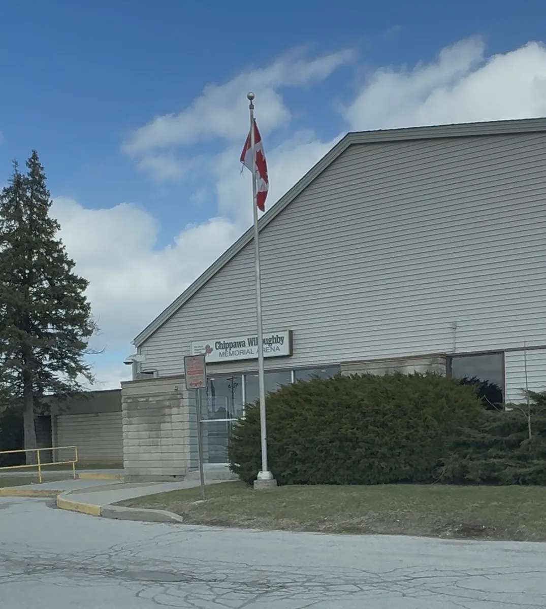 The arena's low front entrance with the Chippawa Willoughby Memorial Arena sign, a flagpole with the Canadian flag and the grey-sided arena roof behind. — Chippawa Willoughby Memorial Arena
