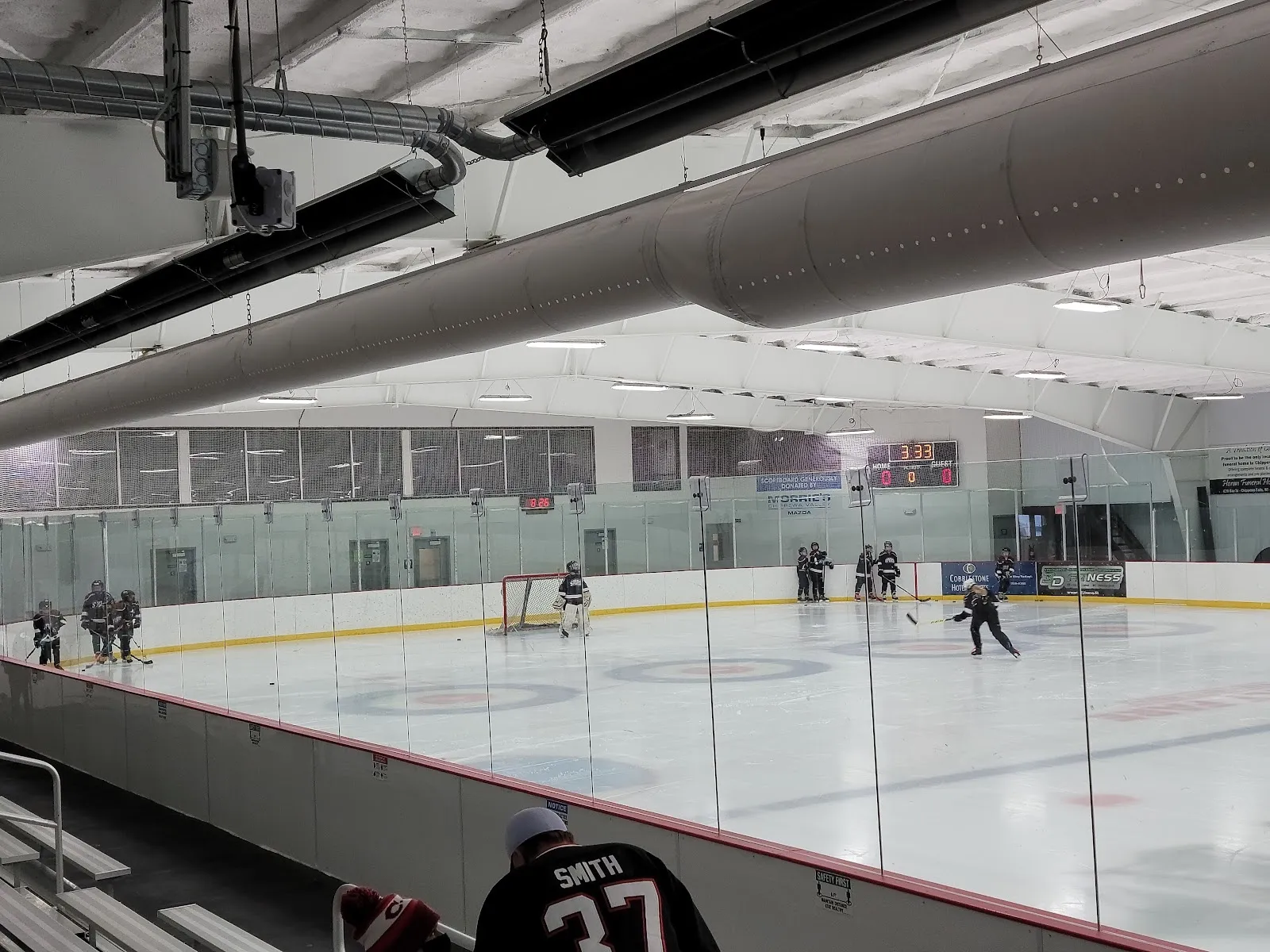 Un partido en curso visto por encima de las vallas, con jugadores repartidos a lo lejos por el hielo bajo un techo claro con un gran ducto de aire y una pared de vidrio verde. — Chippewa Area Ice Arena