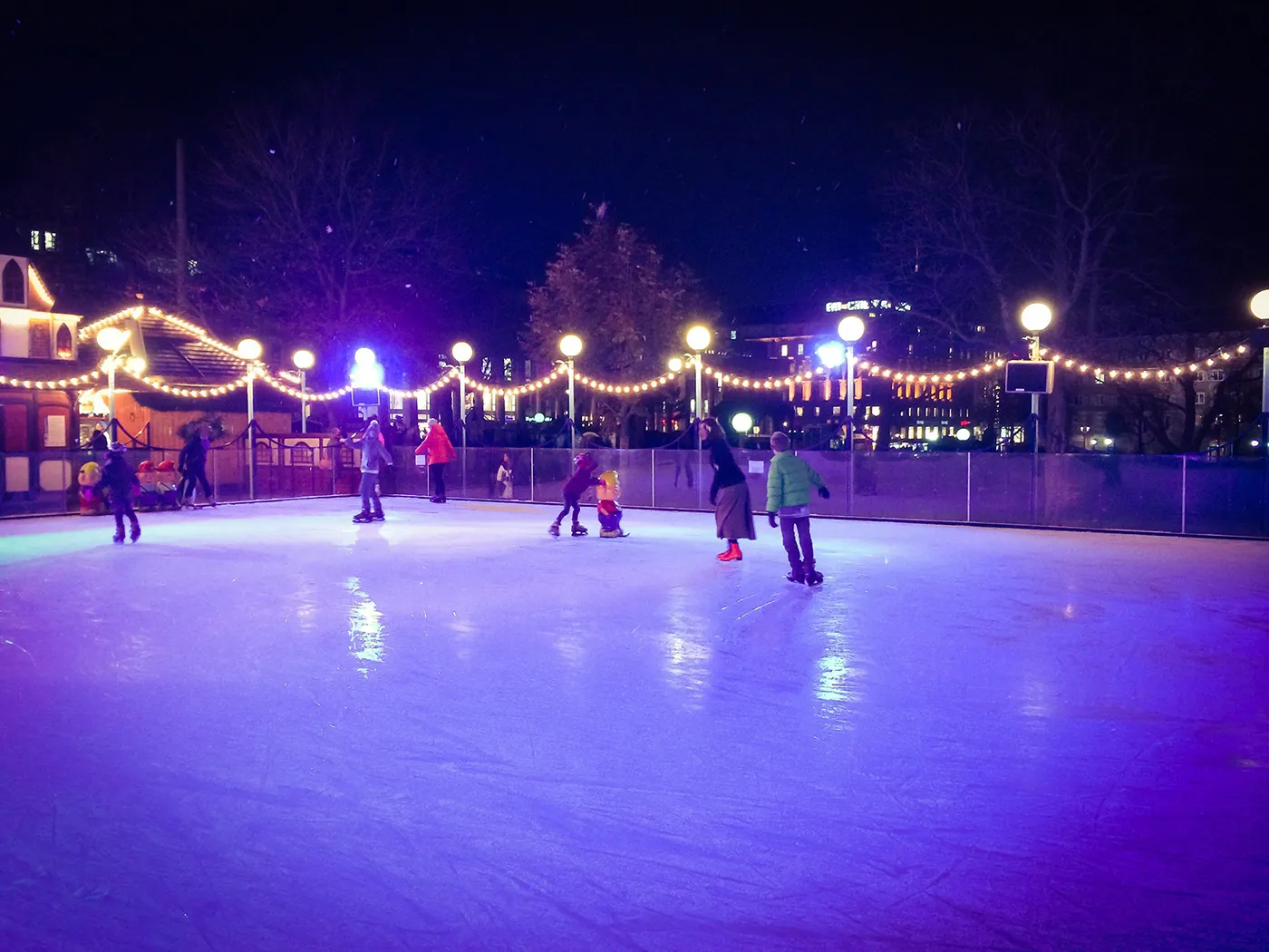 Die beleuchtete Echteis-Eisbahn bei Nacht unter Lichterketten, mit einigen Schlittschuhläufern im Hintergrund. – Eisbahn Christkindlmarkt unterm Maibaum