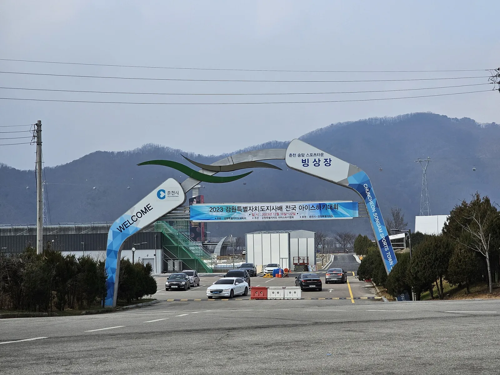 The curved welcome arch at the road entrance to the Songam Sports Town ice rink, with the sign 빙상장 (ice rink) and Chuncheon City's logo, and the grey hall behind. — Chuncheon Songam Sports Town Ice Rink