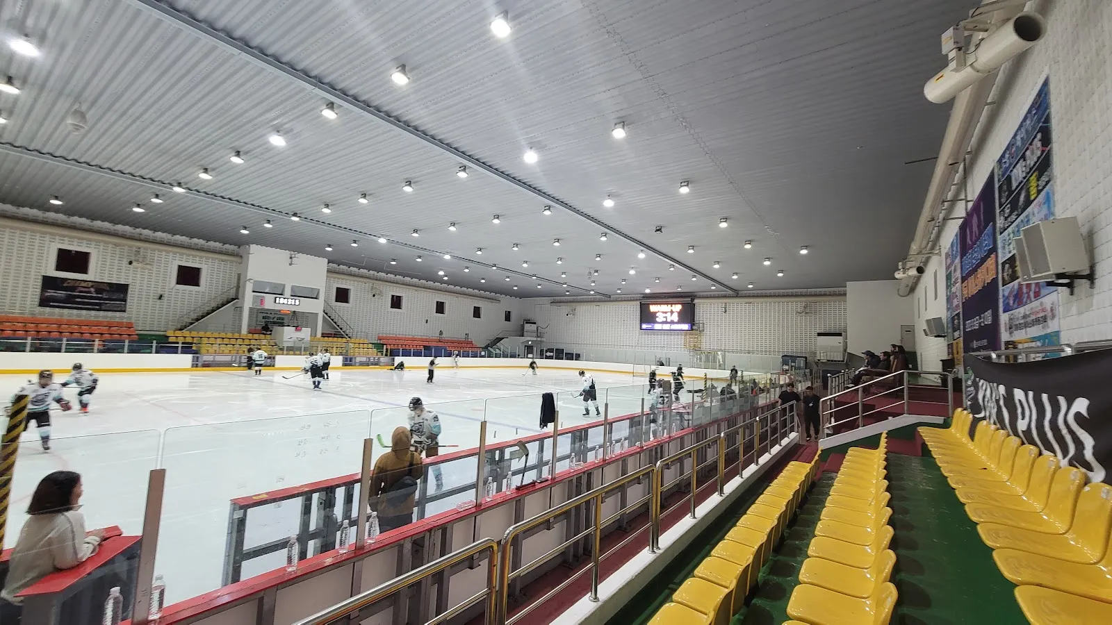 The hall of the Songam Sports Town ice rink from the yellow stand seats, a wide view along the boards to the scoreboard with a few skaters on the ice. — Chuncheon Songam Sports Town Ice Rink