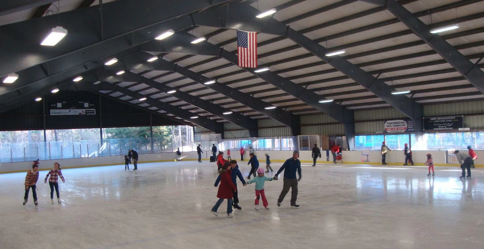 Families on a public skate under the open-sided steel roof, with daylight and trees visible past the far boards. — Churchill Rink at Jackson's Landing