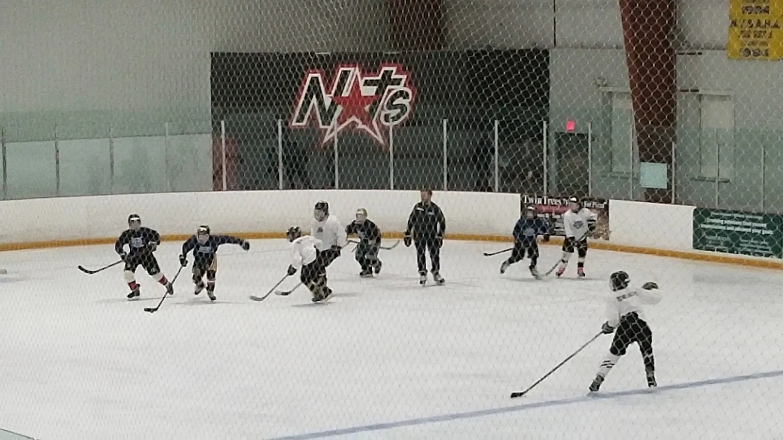 A scrimmage on the ice seen from the boards, a few players spread across the sheet under a wood-clad roof with a red NXT logo on the far netting. — Cicero Twin Rinks
