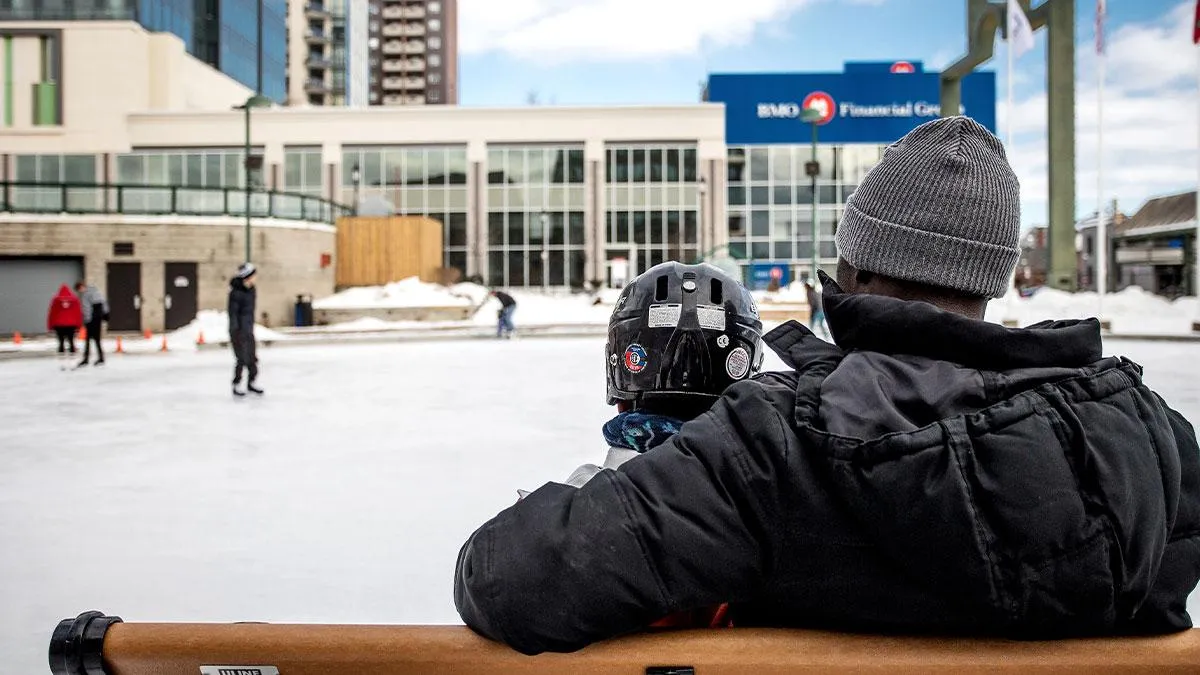 A skater at the edge of the Circle at the Centre rink in Barrie, with the buildings of the city centre behind the ice. — Circle at the Centre Ice Rink (Barrie)