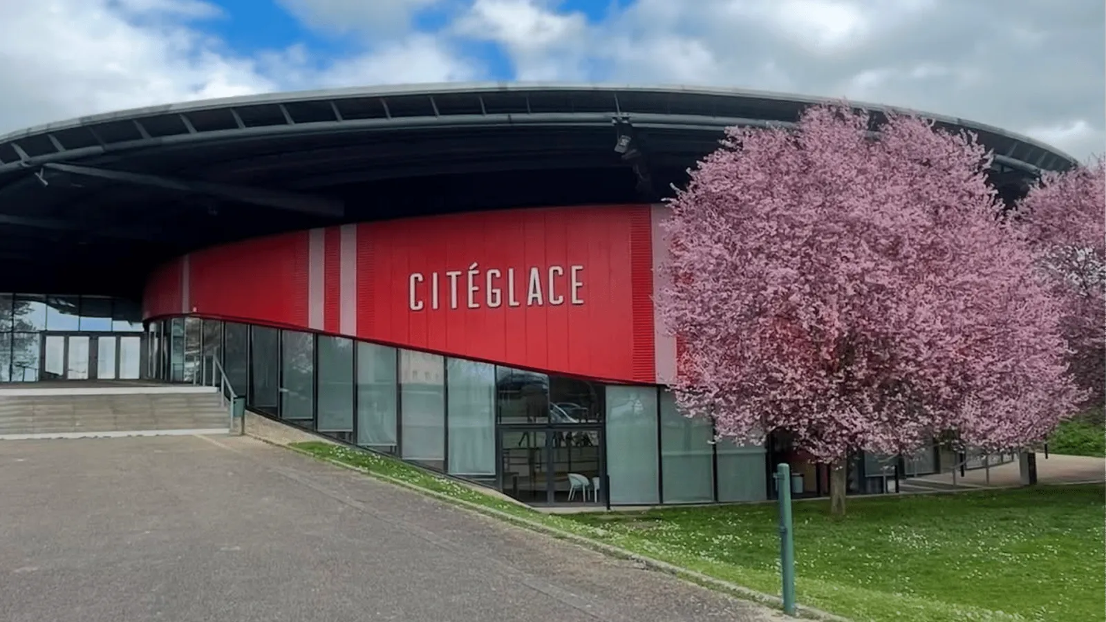 The building's glazed front with a red upper band carrying the CITÉGLACE sign, a flowering tree beside it and a dark curved roof above. — Cité Glace