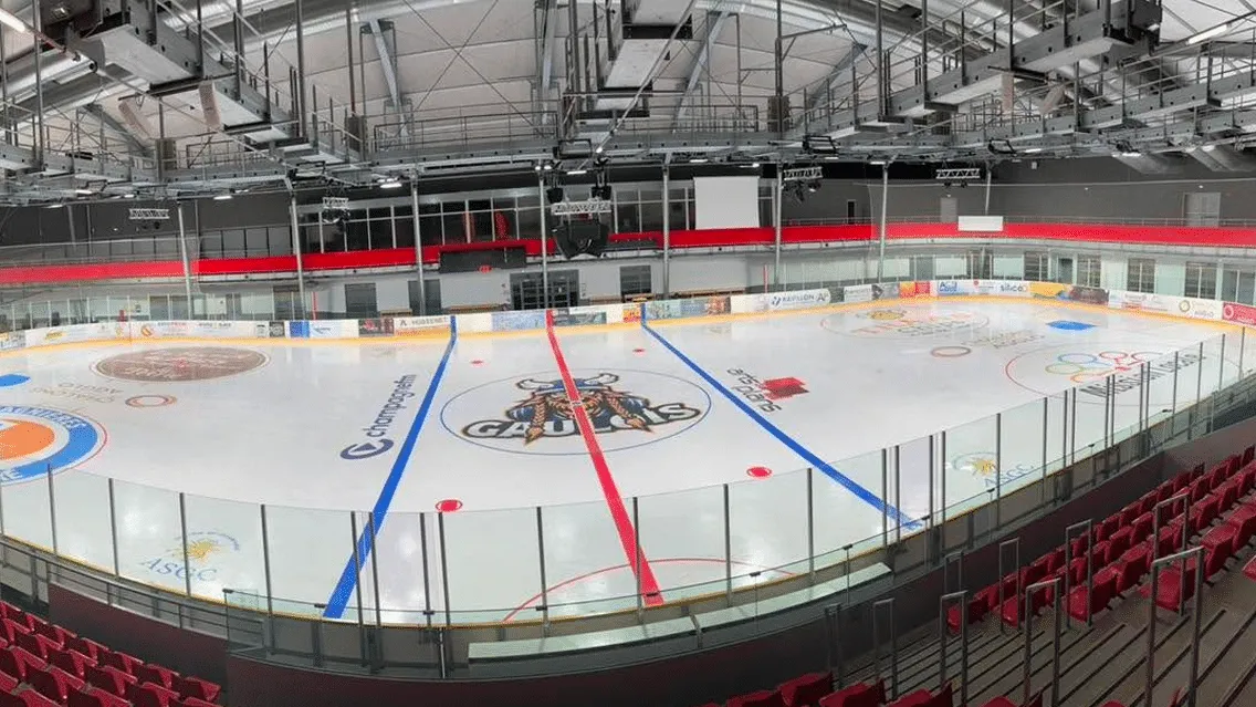 The empty Olympic-size sheet seen from the stands, with a Gaulois logo at centre ice, blue and red lines, red seating along the boards and a steel roof structure. — Cité Glace