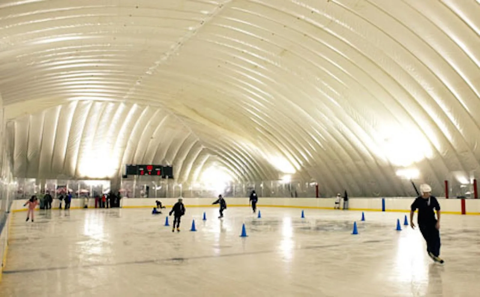 A white inflatable dome arching over the sheet, skaters spread across the ice among blue cones under warm lights. — City Ice Pavilion