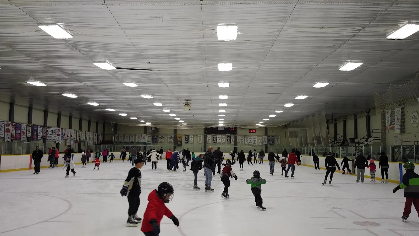 A public skating session spread across a wide sheet, children and adults skating under rows of ceiling lights with banners along the far boards. — Woodstock Civic Centre Arena