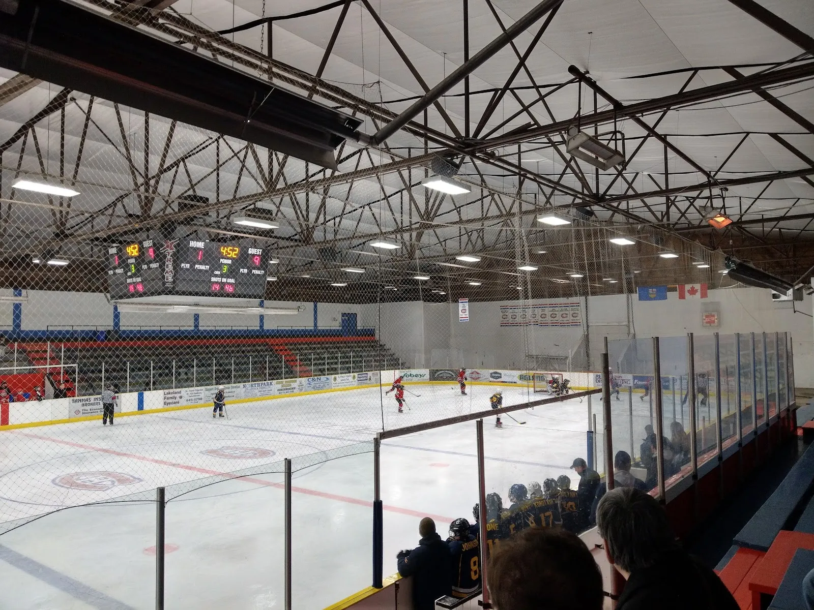 The ice at the Clancy Richard Arena seen from the stands during a hockey game, with the roof trusses, the scoreboard over centre ice and the seating along the far side. — Clancy Richard Arena