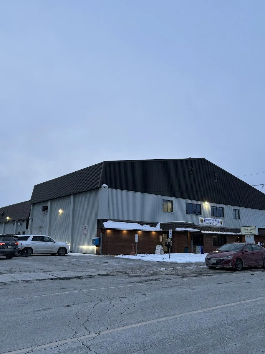 La arena gris con su techo negro y la marquesina iluminada de la entrada, sobre el estacionamiento, al atardecer. — Clarence-Creek Arena