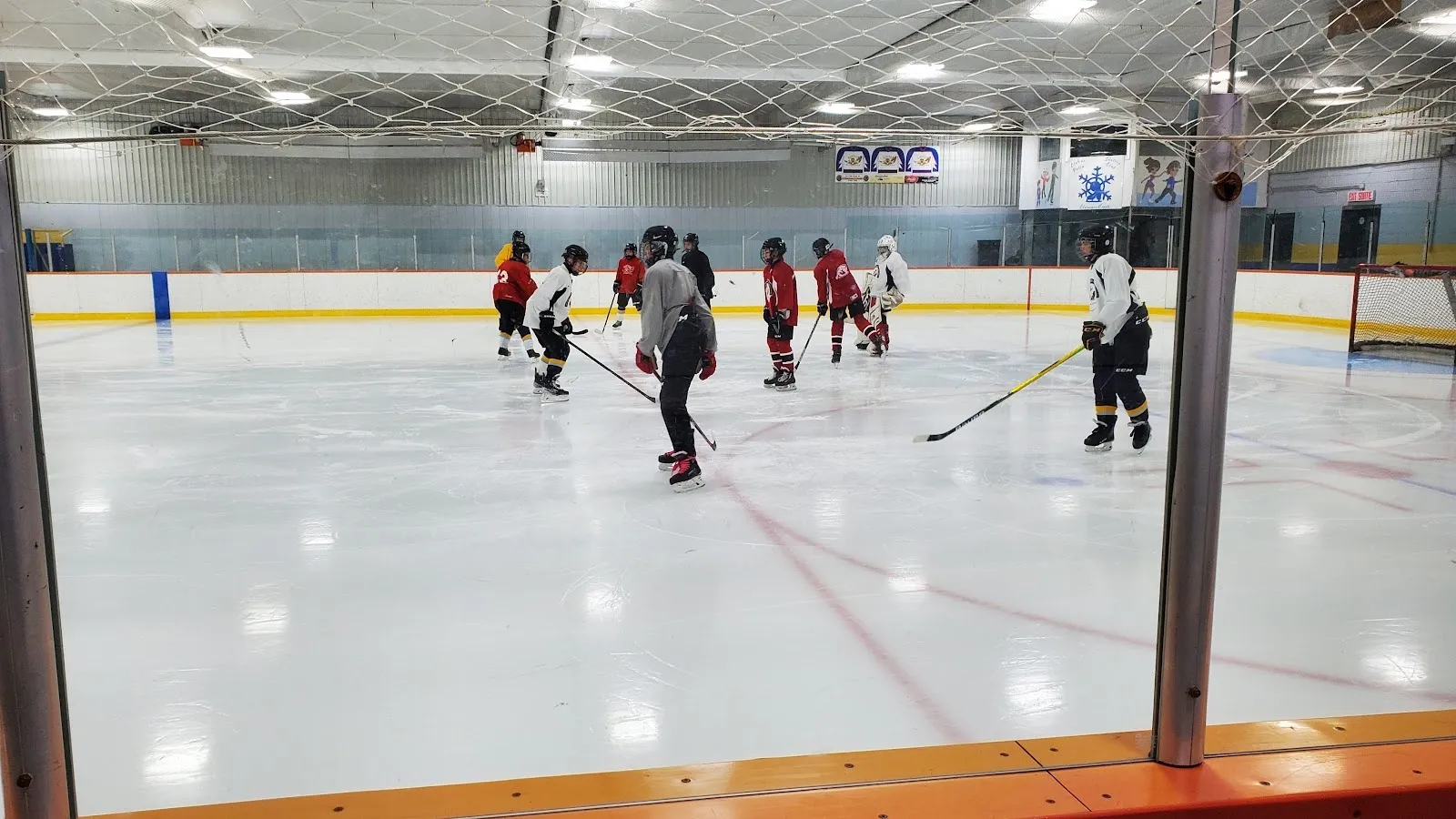 Una práctica en una pista amplia vista a través del vidrio, con jugadores de rojo y blanco repartidos por el hielo bajo un largo techo de luces. — Clarence-Creek Arena
