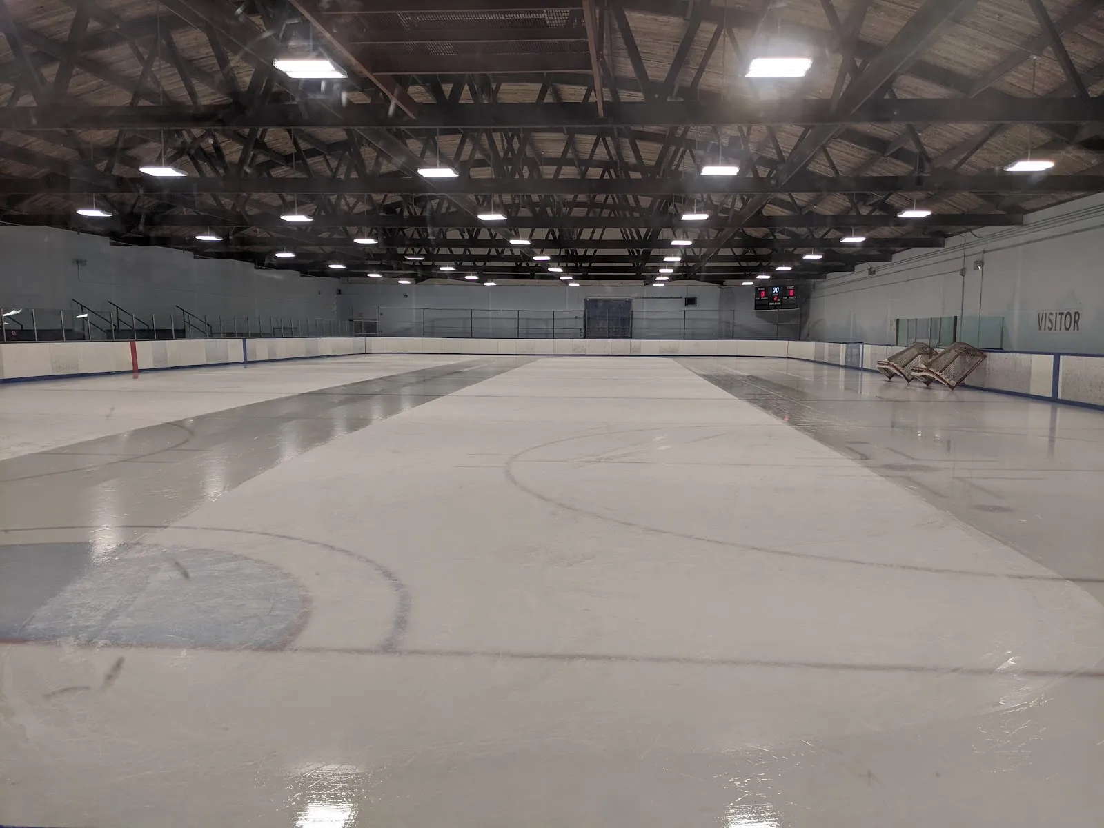 La pista de hielo vacía bajo el oscuro techo de madera, con las luces reflejándose en la superficie recién inundada y el letrero VISITOR en la pared del fondo. — Clarence Mahon Arena