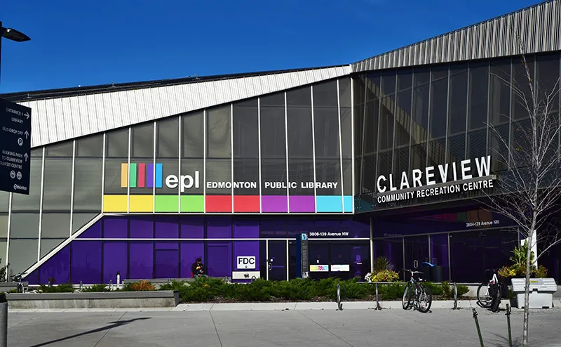 Front entrance of the Clareview Community Recreation Centre building under a clear sky, with large ‘CLAREVIEW COMMUNITY RECREATION CENTRE’ wall signage and Edmonton Public Library branding over the shared glazed facade, bike racks in front. — Clareview Arena