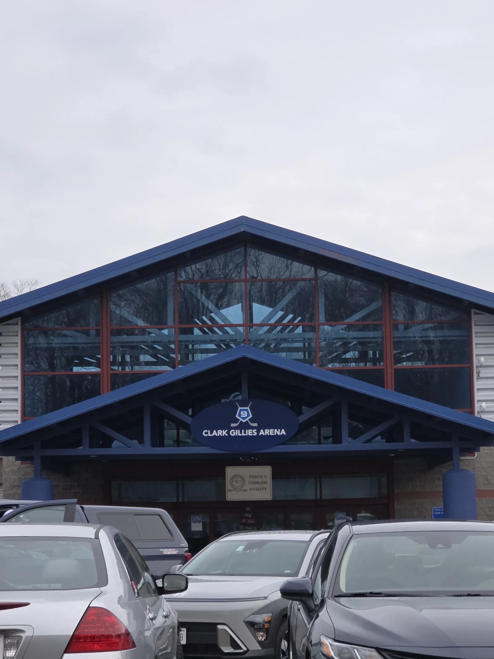 La entrada con frontón de vidrio de la Clark Gillies Arena, con su letrero ovalado del número 9 bajo una línea de techo azul y autos estacionados al frente. — Clark Gillies Arena