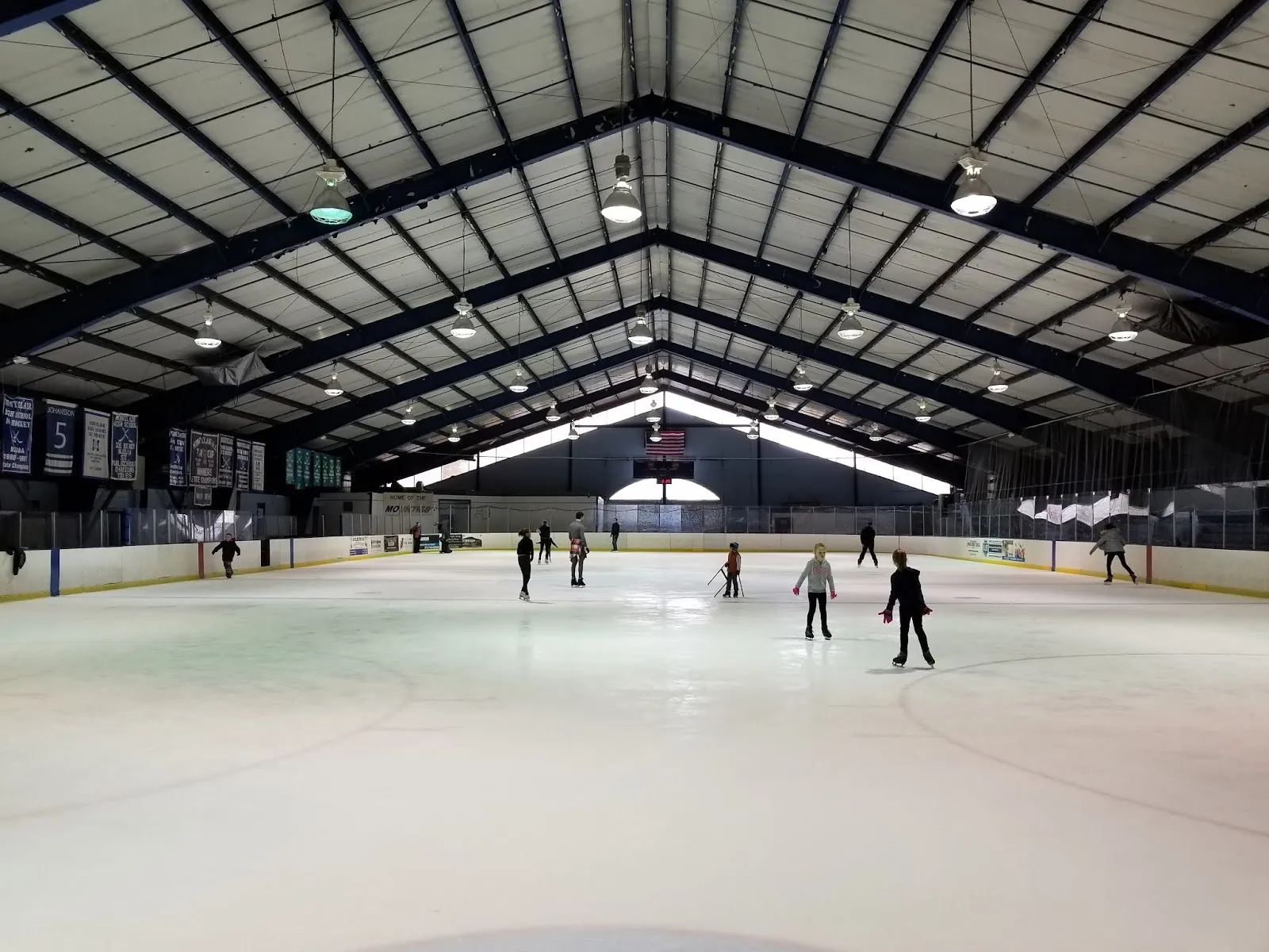 Una vista amplia a lo largo de la pista bajo un techo oscuro de armaduras de acero, con unos cuantos patinadores repartidos por el hielo y banderines del club en la pared del fondo. — Clary Anderson Arena