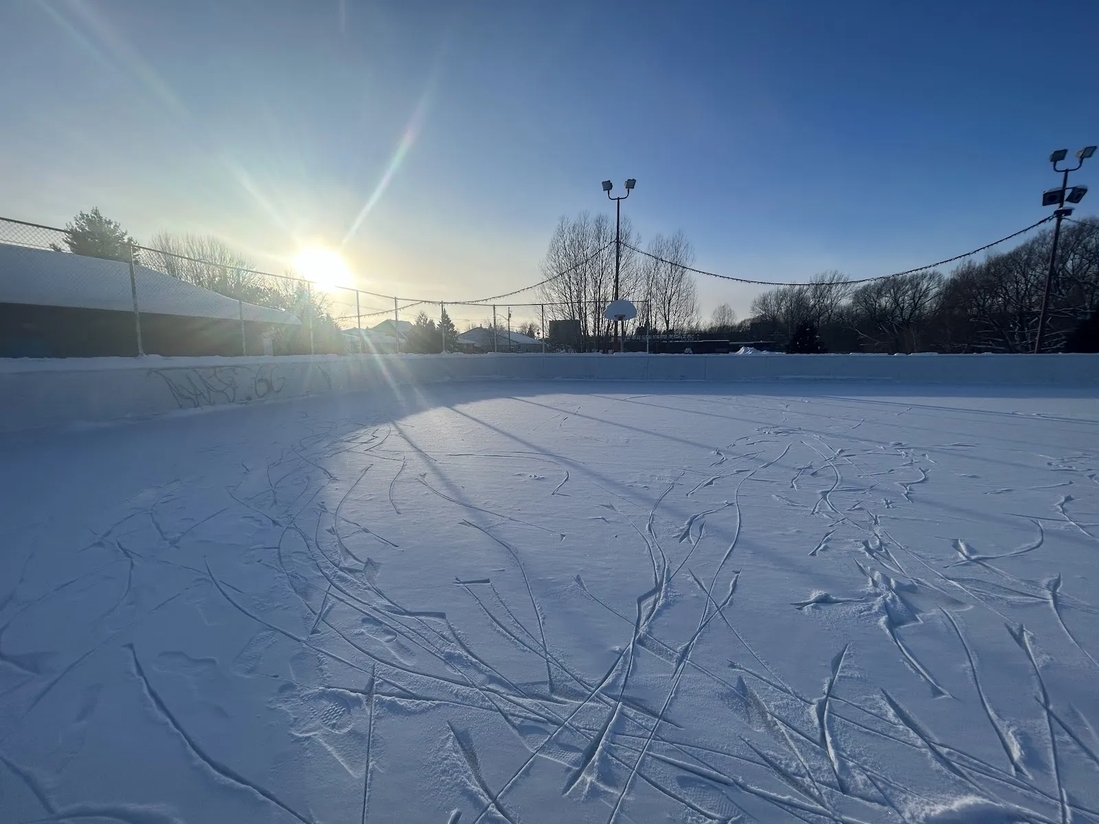 The outdoor rink's ice surface in low winter sun, with skate tracks across the ice and light poles along the far side. — Claude Charbonneau Playground Outdoor Rink